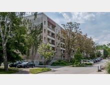 Wohnhaus in einer ruhigen Straße, umgeben von Bäumen und geparkten Autos. Das Gebäude hat mehrere Stockwerke mit Balkonen und eine helle Putzfassade, während der Gehweg von Grünflächen gesäumt ist.