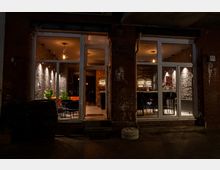 Außenansicht eines modernen Cafés oder einer Bar bei Nacht mit großer Glasfront. Innen sind Tische, Stühle, Pflanzen und eine beleuchtete Theke vor einer mit Stein verkleideten Wand sichtbar.