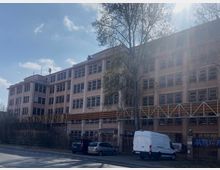 Ein großes vierstöckiges Bürogebäude mit einer Fassade aus beigen rechteckigen Fenstern. Im Vordergrund eine Straße mit parkenden Autos, darunter ein weißer Lieferwagen; daneben ein Baum und ein Gerüst aus Metall und Holz, das Teile des Gebäudes überspannt.