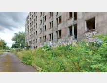 Verlassenes, mehrstöckiges Gebäude mit beschädigten Fenstern und Graffiti auf der Fassade, umgeben von überwucherter Vegetation und einem asphaltierten Weg. Im Hintergrund sind Bäume und ein wolkiger Himmel zu sehen.