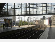 Bahnhof mit erhöhten Bahnsteigen und Eisenbahngleisen, die sich nach rechts biegen. Im Vordergrund ein überdachter Bereich mit Glasfassade, im Hintergrund moderne Gebäude und Oberleitungen.