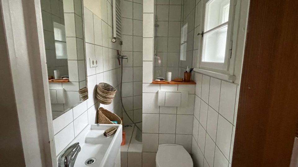 Kleines, schmal geschnittenes Badezimmer mit weißen Wandfliesen, Waschbecken mit Spiegel links und wandhängender Toilette rechts unter einem Fenster mit Milchglas. Am Ende des Raums befindet sich eine geflieste Dusche ohne Kabine, mit Handbrause und Ablageflächen.