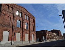 Straßenansicht eines alten, mehrstöckigen Backsteingebäudes mit verzierten Fensterbögen und teilweise zugemauerten Eingängen. Daneben erstrecken sich weitere ähnliche Gebäude entlang einer gepflasterten Straße unter klarem, blauem Himmel.