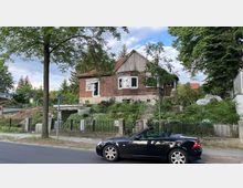 Einstöckiges Haus mit unvollständiger, teils hölzerner Fassade und rotem Ziegeldach, umgeben von einem verwilderten Garten. Im Vordergrund stehen ein Baum, ein schwarzes Cabriolet, ein Parkverbotsschild und ein niedriger Zaun entlang einer Asphaltstraße.