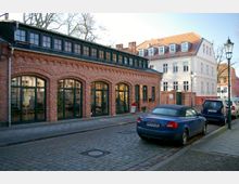 Backsteingebäude mit großen, bogenförmigen Fenstern und dunkelgrünen Rahmen, davor eine gepflasterte Straße mit geparkten Autos; im Hintergrund ein weiß verputztes Mehrfamilienhaus mit Satteldach.