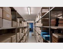 Ein Archivraum mit langen Metallregalen, die mit beschrifteten Kartons, Ordnern und gebundenen Dokumenten gefüllt sind. Eine Leuchtstoffröhre an der Decke beleuchtet den schmalen Gang zwischen den Regalen, und im Hintergrund ist ein kleines Fenster zu sehen.