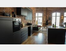 Offene Loftwohnung mit Backsteinwänden, bestehend aus einer modernen Küche mit schwarzen Schränken und Geräten sowie einem Wohnbereich mit Holzfußboden, einem grünen Sofa und großen, bogenförmigen Fenstern, die viel Tageslicht hereinlassen. Im Raum stehen ein Esstisch und dekorative Elemente wie Pflanzen und Lampen.