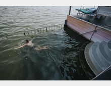 Ein Mann schwimmt in einem Gewässer neben einer Holzplattform mit Metallgeländern und Liegestühlen; daneben führen gebogene Stufen ins Wasser.