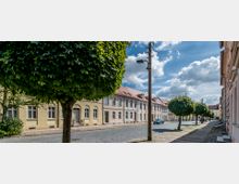 Gepflasterte Straße in einer ruhigen Wohngegend mit Reihenhäusern im klassischen Stil und roten Ziegeldächern. Links und rechts der Straße stehen regelmäßig gepflanzte Bäume, begleitet von einem Gehweg mit kleinen Grünflächen; der Himmel ist teils bewölkt.