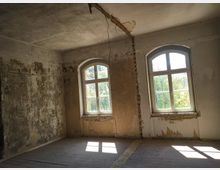 Ein leerer, verfallener Raum mit abblätterndem Putz an den Wänden, zwei großen bogenförmigen Fenstern, durch die Tageslicht einfällt, und einem Holzboden. Außen ist grüne Vegetation sichtbar.