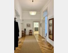 Heller Flur mit Holzboden und einem beigen Läufer, ausgestattet mit Holzmöbeln, einem großen Spiegel an der rechten Wand und weißen Wänden, die mit dezenten Kunstwerken geschmückt sind. Im Hintergrund führen geöffnete Türen in andere helle Räume mit ähnliche Einrichtung.