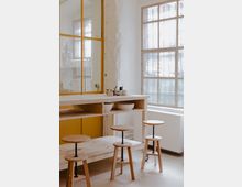 Ein minimalistischer Innenraum mit einer weißen Ziegelwand, großen Sprossenfenstern, einem rechteckigen Holztisch mit offenen Regalfächern und mehreren schlichten Holzstühlen mit runden Sitzflächen. Im Hintergrund befinden sich gelbe Schranktüren und ein Spiegel mit gelbem Rahmen.