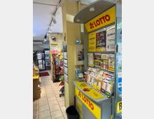 Ein kleiner Laden mit einem Lotto-Schalter im Vordergrund, einschließlich Prospekten und Wettscheinen auf einem gelben Tresen mit der Aufschrift "LOTTO". Im Hintergrund sind Regale mit Getränken, Zeitschriften und Snacks zu sehen; der Boden ist mit hellen Fliesen ausgelegt.