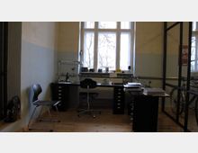 Ein Arbeitszimmer mit einem großen Fenster, das Tageslicht hereinlässt. Der Raum enthält zwei schwarze Arbeitstische, einen Schreibtischstuhl, einen zusätzlichen Stuhl und verschiedene Gegenstände wie einen Schreibtischlampenarm, Bücher und Bürogeräte. Der Holzboden und eine Glas-/Metalltrennwand definieren den Raum weiter.