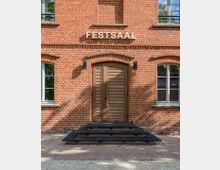 Backsteinfassade eines Gebäudes mit der Aufschrift "FESTSAAL". Zentrale Holztür mit Rundbogen, flankiert von zwei Fenstern und einer kleinen Metallrampe davor.