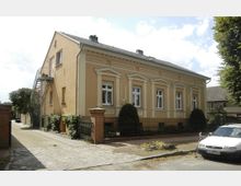 Gelbes zweistöckiges Wohnhaus mit historischer Fassade und weißen Fensterrahmen, verziert mit Säulen und Fassadenelementen. Davor steht ein weißes Auto, und das Haus ist von einem schmiedeeisernen Zaun und kleinen Bäumen umgeben.