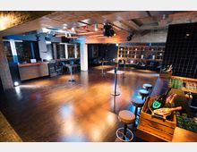 Ein moderner Veranstaltungsraum mit einer großen Tanzfläche aus Holz, einer Bar mit Hockern auf der rechten Seite und hohen Stehtischen. Im Hintergrund befinden sich Sitzgelegenheiten, Regale mit Deko-Objekten und große Fenster mit Blick auf eine Stadt bei Nacht.