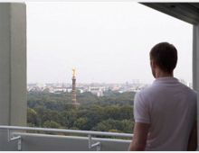 Ein Mann steht auf einem Balkon und blickt über einen Park mit dichtem Baumbestand, in dessen Mitte die Berliner Siegessäule mit der goldenen Figur sichtbar ist. Im Hintergrund erstreckt sich die Skyline von Berlin mit weiteren Gebäuden.