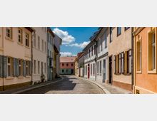 Gepflasterte, enge Gasse mit historischen Wohnhäusern in warmen Farbtönen wie Beige, Weiß und Orange; die Fassaden sind mit Fensterläden, teilweise geöffneten Fenstern und Blumentöpfen dekoriert, im Hintergrund ein Haus mit roten Ziegeldächern und blauem Himmel.
