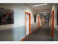 Langer Flur in einem modernen Gebäude mit grauem Boden und Wänden, die zur Hälfte weiß und grau gestrichen sind. Türen mit orangefarbenen Türrahmen und Wandbilder prägen die Gestaltung; am Ende des Flurs ist eine geöffnete Tür zu sehen.