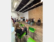 Interior of an open studio-style room with wood floors, a long exposed brick wall, and a grid ceiling with acoustic panels and hanging lights. Bright green chairs are arranged in rows facing a small setup at the front, with a pride flag and wall-mounted deer head decoration on the far wall.