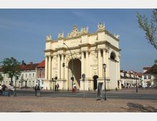 Monumentales Stadttor in klassizistischer Architektur mit reich verzierten Säulen, Skulpturen und Reliefs, umgeben von historischen Gebäuden. Im Vordergrund sind eine gepflasterte Straße, Fußgänger und Straßenlaternen zu sehen.