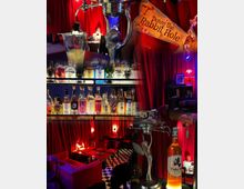 Eine Bar mit gemütlicher Atmosphäre, dekoriert mit roten Vorhängen und gedämpfter Beleuchtung. Im Vordergrund stehen eine Auswahl an Spirituosenflaschen, eine Absinthe-Fontäne und ein Schild mit der Aufschrift „Down the Rabbit Hole“, im Hintergrund befinden sich Sitzbereiche mit roten Polstern und ein schwarz-weiß karierter Boden.