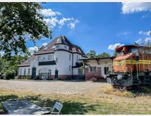 Ein weißes Gebäude mit rotem Ziegelsockel und einem markanten Turmdach, gelegen an einem gepflasterten Hof. Rechts im Bild steht eine alte, rot-gelb lackierte Lokomotive; im Vordergrund eine Sitzbank unter einem Baum, umgeben von Gras und Bäumen.