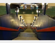 Treppenabgang einer U-Bahn-Station mit blauen Seitenwänden, roten Handläufen und modernem Beleuchtungssystem. Im Hintergrund sind grün geflieste Wände und bogenförmige Betonstrukturen zu sehen, die den unterirdischen Raum definieren.