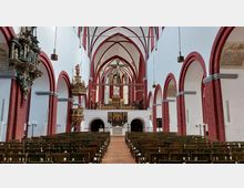Innenraum einer historischen Kirche mit gotischen Spitzbögen, roten Backsteinarkaden und weiß verputzten Wänden. Im Vordergrund stehen Reihen von Holzbänken, während der Blick auf den reich verzierten Altar im Hintergrund gerichtet ist, flankiert von einer Kanzel und bunten Glasfenstern.