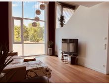 Modernes Wohnzimmer mit großen, deckenhohen Fenstern, die einen Blick auf grüne Bäume bieten. Der Raum ist mit einem Sofa, einem Fernseher auf einem kleinen Tisch und Holzfußboden eingerichtet, ergänzt durch Zimmerpflanzen und eine minimalistische Dekoration.