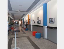 Langer, moderner Eingangsbereich mit hellen Wänden, bunten Säulen in Blau und Orange sowie Dekorationen wie Bilderrahmen an den Wänden. Der Raum ist mit Stehtischen, bunten Würfelhockern und schwarzen Ablageflächen ausgestattet; im Hintergrund befindet sich eine Rezeption.