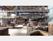 Innenansicht einer stillgelegten Industrieanlage mit rostigen Metallstrukturen, großen Gießbehältern und schweren Maschinen. Im Hintergrund sind Stahlträger, Gestelle und Lichter zu sehen, die auf eine ehemalige Nutzung im Schwerindustriebereich hinweisen.