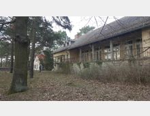 Ein großes, gelbliches, zweistöckiges Gebäude mit schrägem Ziegeldach und einer Terrasse mit Säulen im Vordergrund, umgeben von Bäumen und braunem Laubboden in einer ländlichen, bewachsenen Umgebung. Im Hintergrund ist ein weiteres kleines Gebäude mit rotem Dach sichtbar.