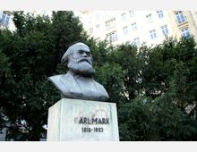 Büste von Karl Marx auf einem Sockel mit der Inschrift „Karl Marx 1818–1883“, umgeben von dichten grünen Bäumen im Vordergrund; im Hintergrund ein mehrstöckiges Gebäude mit hellen Fassaden und zahlreichen Fenstern.