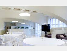 Moderner Veranstaltungsraum mit weißen runden Tischen und weißen Stühlen, großen Fenstern, bogenförmigen Decken und Hängeleuchten. Im Hintergrund sind Pflanzen auf einer erhöhten Ebene und ein großer schwarzer Bildschirm auf einem Ständer erkennbar.