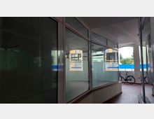 Ein überdachter Durchgang mit großen Glasfenstern auf der rechten Seite, die Büroräume mit transparenten und teilweise matten Scheiben zeigen. Im Hintergrund sind Fahrräder, ein offener Außenbereich und ein mehrstöckiges Gebäude sichtbar.