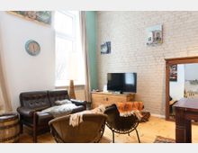 Wohnzimmer mit gemauerter Ziegelwand, ausgestattet mit einem schwarzen Ledersofa, zwei Sesseln mit orangefarbenen Decken, einem Holztisch und einem niedrigen Schrank, der als Fernsehmöbel dient. An der Wand hängen gerahmte Bilder, und ein großes Fenster lässt natürliches Licht in den Raum.