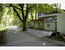 Ein grüner Parkweg mit großen Bäumen im Vordergrund, daneben ein kleines Backsteingebäude mit grünen Türen und Fensterläden. Im Hintergrund ist ein modernes Gebäude mit Glasfassade zu erkennen.