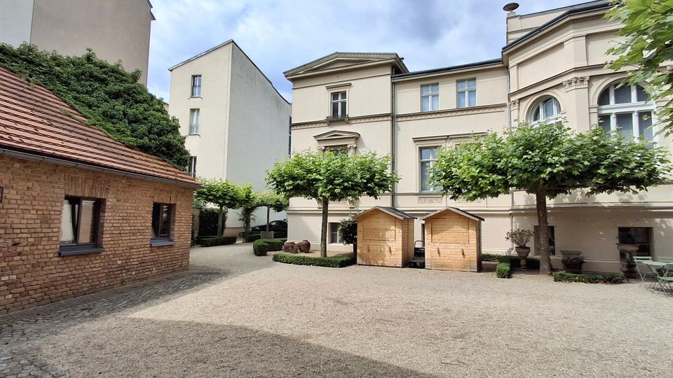 Innenhof zwischen historischen Gebäuden mit heller Putzfassade und großen Rundbogenfenstern sowie einem niedrigen Backsteinhaus mit Ziegeldach. Auf dem geschotterten Platz stehen beschnittene Bäume, zwei kleine Holzhütten und Sitzmöbel mit geschlossenem Sonnenschirm.
