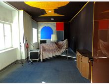 Ein einfacher Innenraum mit dunklen Wänden und einer gelben Decke mit dekorativen Mustern. Im Raum befinden sich ein überzogener Tisch, eine Lampe, Aufnahmeausrüstung, ein Fenster mit Tageslicht auf der linken Seite und eine blaue Fläche in einem gemauerten Bogen an der gegenüberliegenden Wand.