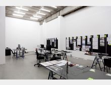 Offener Büro- oder Arbeitsbereich mit hohen Decken und Sichtbetonbalken; ausgestattet mit mehreren Glastischen, Monitoren, Stühlen und verstreuten Arbeitsmaterialien. Eine Wand ist mit schwarzen Pinnwänden bedeckt, auf denen Zeichnungen, Skizzen und Notizen angebracht sind.