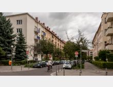 Wohnstraße in einer Stadt mit mehrstöckigen, cremefarbenen Wohngebäuden mit Balkonen. Entlang der Straße stehen geparkte Autos, Bäume und Straßenlaternen bei bedecktem Himmel, während ein Fahrradfahrer durch die Szene fährt.