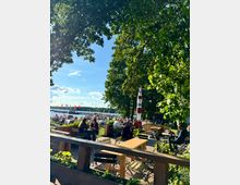 Ein Biergarten am Ufer eines Sees mit Blick auf Segelboote und rote Flaggen; im Vordergrund stehen Holztische und Stühle unter Bäumen, neben einem dekorativen rot-weißen Leuchtturm.