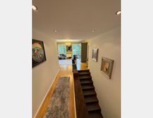 Innenansicht eines Wohnbereichs mit Blick auf ein helles Wohnzimmer mit großen Fenstern, die auf eine grüne Außenansicht blicken. Im Vordergrund befinden sich eine Treppe mit dunklen Stufen, ein Holzfußboden und dekorative Bilder an den weißen Wänden.