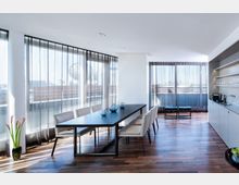 Heller und moderner Ess- und Wohnbereich mit großen, bodentiefen Fenstern, die von transparenten Vorhängen bedeckt sind. Der Raum hat einen langen Holztisch mit weißen Stühlen, Holzboden und eine Küchenzeile mit offenen Regalen auf der rechten Seite.