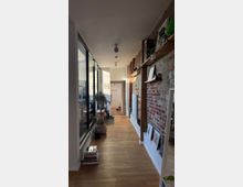 Schmaler Flur in einem modernen Innenraum mit einer roten Backsteinmauer auf der rechten Seite und einer großen Fensterfront auf der linken Seite. Der Boden besteht aus hellem Holz, und Regale sowie dekorative Pflanzen sind entlang der Backsteinmauer platziert.