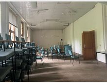 Ein leerer, einfacher Raum mit weißen Wänden und einer abgehängten Decke. Stühle mit blauen Polstern sind teils gestapelt, teils ordentlich aufgestellt; eine Holztür steht offen, und mehrere große Fenster lassen Tageslicht herein.