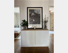 Weiße Kommode mit Holzplatte in einem Wohnraum, darauf stehen eine Kerze, ein gerahmtes Schwarz-Weiß-Foto, ein Topf mit einer Zimmerpflanze und ein Strauß mit Zweigen in einer Vase. Im Hintergrund sind Holzböden, ein Bett und ein abgetrennter Wohnbereich zu sehen.