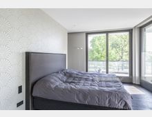 Helles Schlafzimmer mit einem großen Bett, gemusterten Tapeten und bodentiefen Fenstern, die auf einen Balkon mit Blick auf grüne Bäume führen.